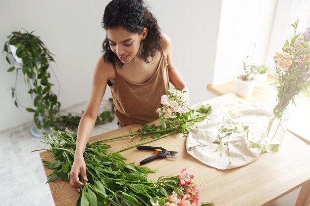 Young beautiful african woman florist taking care of flowers at workplace over white wall.