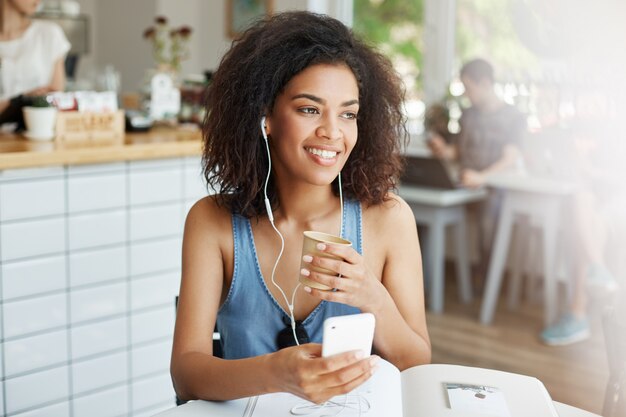 Young beautiful african woman student listening to music in headphones smiling sitting at table with books at cafe.