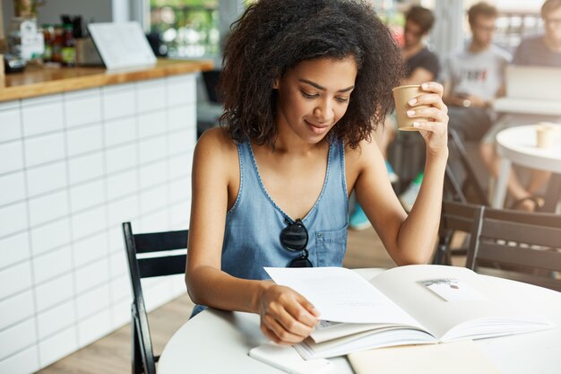 コーヒーを飲みながら雑誌を見て笑ってカフェに座っている若い美しいアフリカ女性学生。学習と教育。