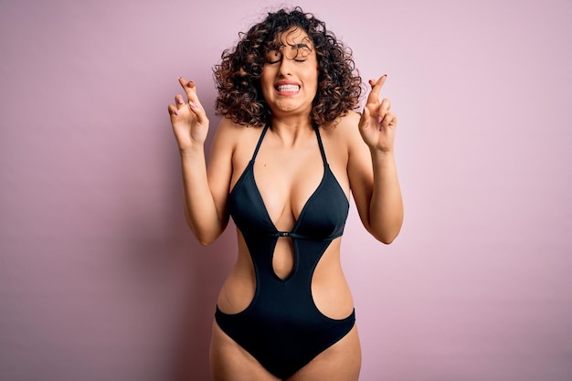 Young beautiful arab woman on vacation wearing swimsuit and sunglasses over pink background gesturing finger crossed smiling with hope and eyes closed Luck and superstitious concept