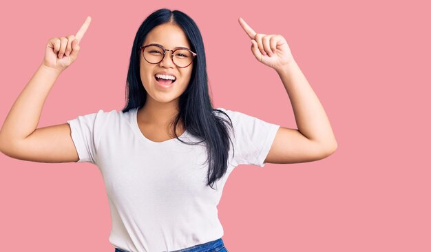 Young beautiful asian girl wearing casual clothes and glasses smiling amazed and surprised and pointing up with fingers and raised arms.