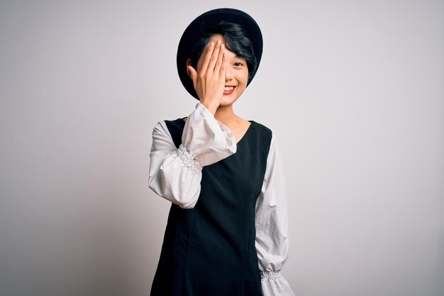 Young beautiful asian girl wearing casual dress and hat standing over isolated white background covering one eye with hand confident smile on face and surprise emotion