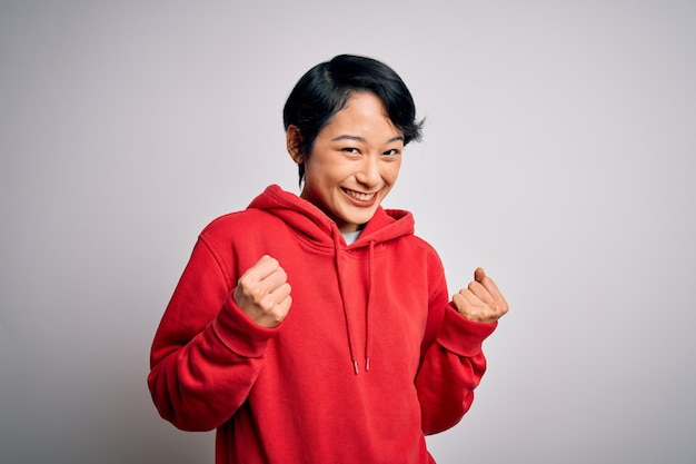 Young beautiful asian girl wearing casual sweatshirt with hoodie over white background excited for success with arms raised and eyes closed celebrating victory smiling Winner concept