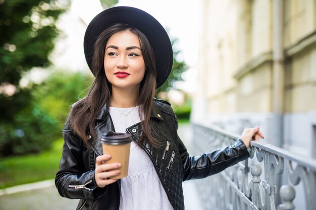 Young beautiful Asian girl with a paper cup of coffee walking through the summer city