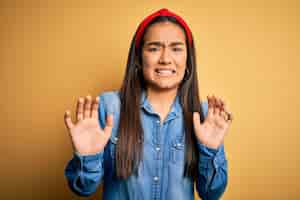 Free photo young beautiful asian woman wearing casual denim shirt and diadem over yellow background disgusted expression displeased and fearful doing disgust face because aversion reaction with hands raised