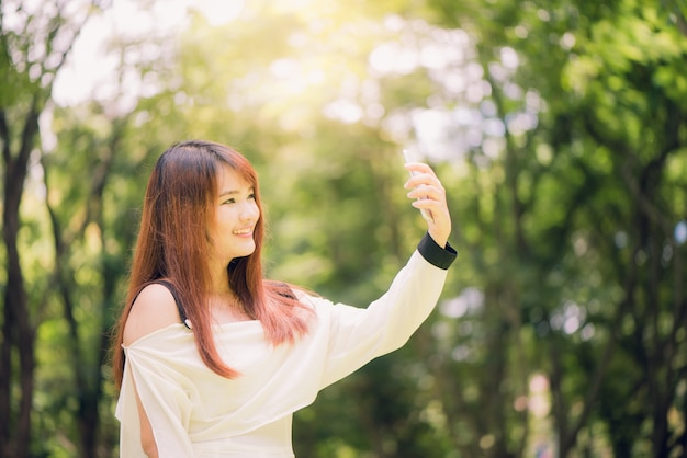 공원에서 그녀의 전화에는 selfie를 복용하는 긴 갈색 머리를 가진 젊은 아름 다운 아시아 여자. 자연 조명, 생생한 색상.