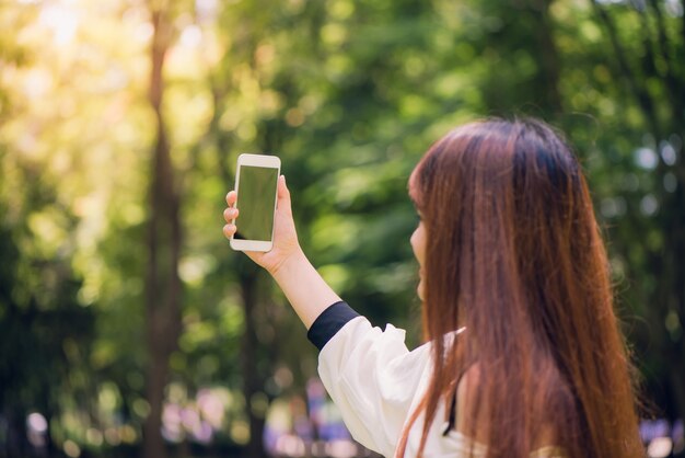 長い茶色の髪を持つ若い美しいアジアの女性は、公園内の彼女の電話でセルフリーを取っています。自然光、鮮やかな色。