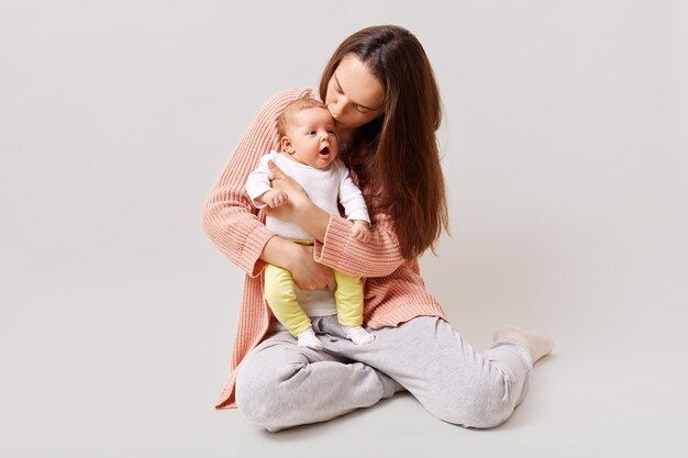 Young beautiful attractive mother holding and kissing newborn baby while sitting on floor