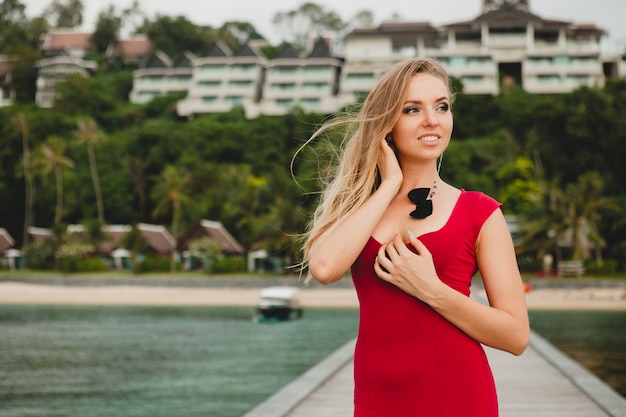 Young beautiful attractive woman standing alone on pier in luxury resort hotel, summer vacation, red long dress, blond hair, sexy apparel, tropical beach, seductive, sensual, smiling