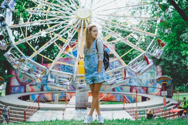 молодая красивая блондинка девушка в джинсовой целом с рюкзаком, создавая в парке развлечений