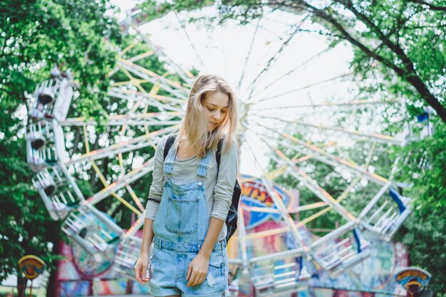 young beautiful blond girl in denim overall with a backpack posing in an amusement park