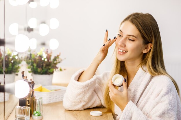 Young beautiful blonde girl cares for face skin with moisturizer in front of mirror