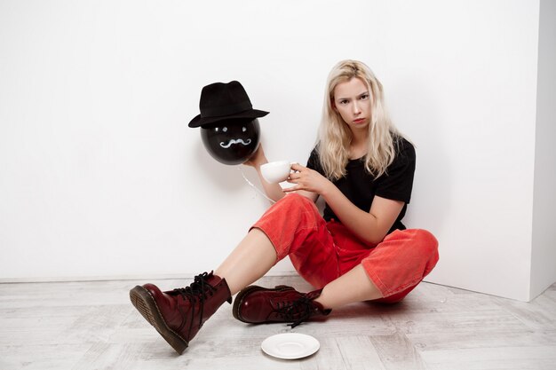 Young beautiful blonde girl holding black baloon in hat sitting on floor drinking coffee over white wall.