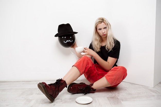 白い壁にコーヒーを飲みながら床に座って帽子に黒い風船を保持している美しい金髪少女。