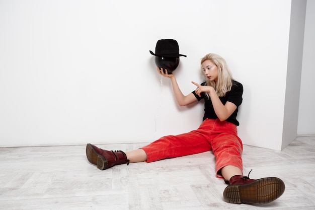 Young beautiful blonde girl holding black baloon in hat sitting on floor on white wall. Copy space