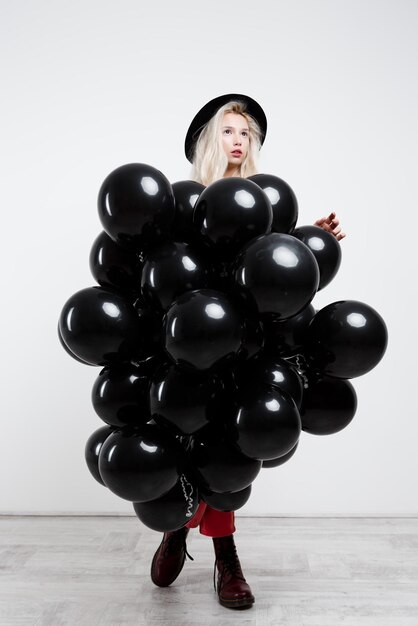 Young beautiful blonde girl holding black baloons over white wall