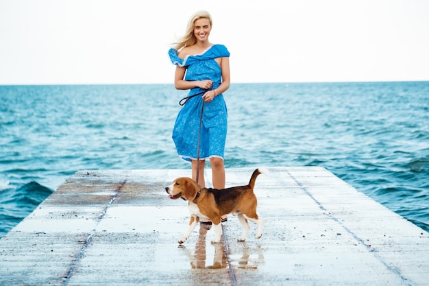 Giovane bella ragazza bionda che cammina, giocando con il cane da lepre in riva al mare.