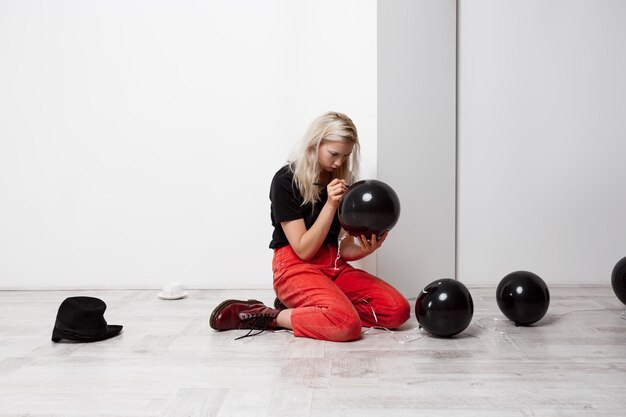 Young beautiful blonde girl with black baloon sitting on floor over white wall