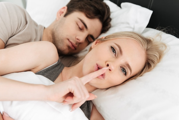 Молодая красивая блондинка показывает жест молчания, лежа в постели со своим мужчиной