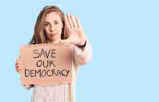 Foto gratuita giovane bella donna bionda che tiene salva la nostra bandiera della democrazia con la mano aperta che fa il segnale di stop con un gesto di difesa dell'espressione seria e sicura