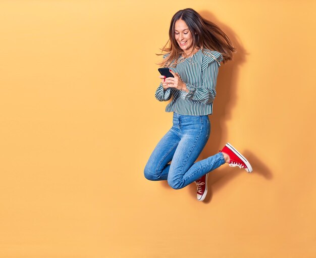 Young beautiful blonde woman jumping with smile on face. Using smartphone over isolated yellow background