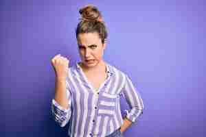 Free photo young beautiful blonde woman wearing casual striped shirt standing over purple background angry and mad raising fist frustrated and furious while shouting with anger rage and aggressive concept