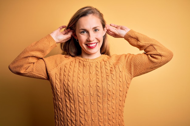 Foto gratuita giovane bella donna bionda che indossa un maglione casual in piedi su sfondo giallo sorridente tirando le orecchie con le dita gesto divertente problema di audizione