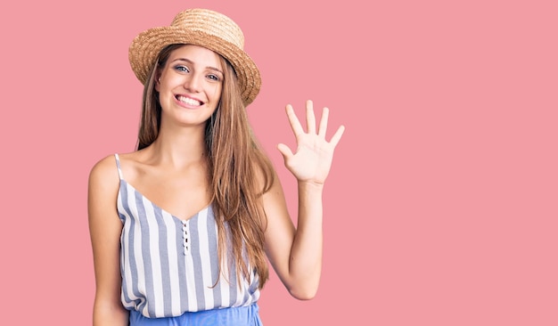 Foto gratuita giovane bella donna bionda che indossa un cappello estivo che mostra e punta verso l'alto con le dita numero cinque mentre sorride fiducioso e felice.