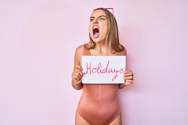 Young beautiful blonde woman wearing swimwear and holding holidays banner angry and mad screaming frustrated and furious, shouting with anger. rage and aggressive concept.