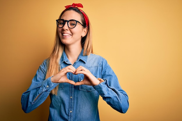 Free photo young beautiful blonde woman with blue eyes wearing denim shirt over yellow background smiling in love showing heart symbol and shape with hands romantic concept