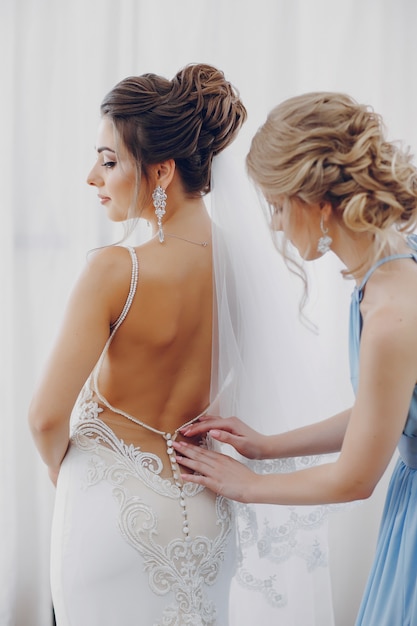 A young and beautiful bride at home standing near window with bridesmaid