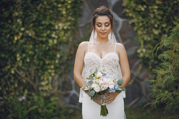 A young and beautiful bride is standing in a summer park with bouquet of flowers