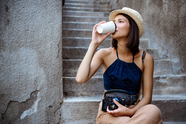 Молодая красивая брюнетка женский фотограф, пить кофе, сидя на лестнице.