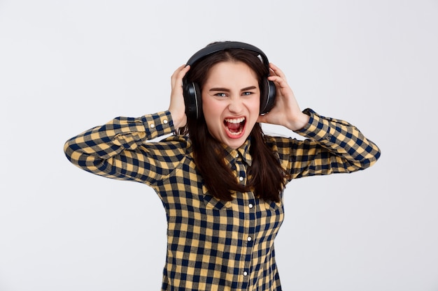 Young beautiful brunette girl dressed in plaid shirt listening music in headphones screaming