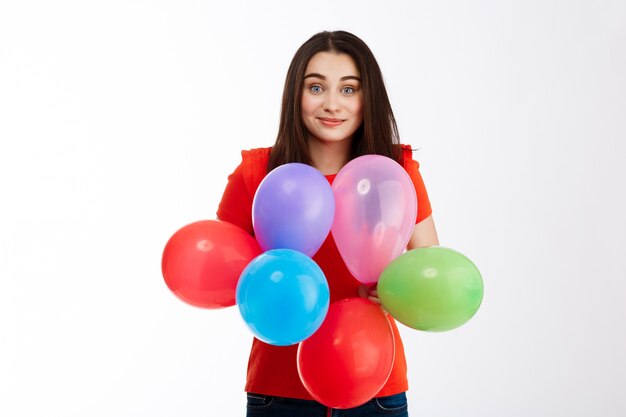 Young beautiful brunette girl dressed in red holding colorful baloons smiling over white wall