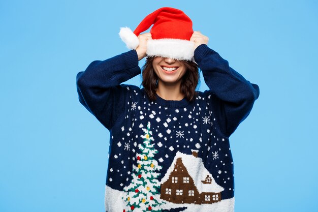 Молодая красивая девушка брюнетка в вязаный свитер и Рождество шляпу, улыбаясь через синюю стену