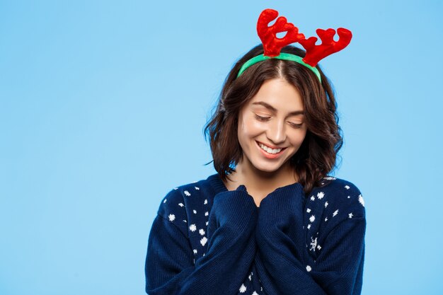 青い壁に笑みを浮かべてニットセーターとクリスマスのトナカイの枝角の若い美しいブルネットの少女