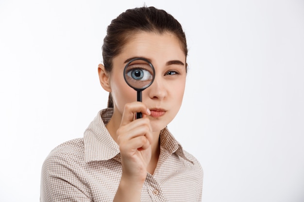 Young beautiful brunette girl looking with mistrust at camera through magnifier over white wall.