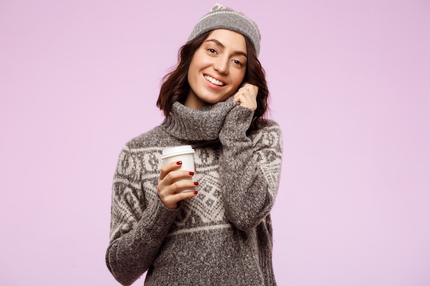 Young beautiful brunette girl smiling holding coffee over light wall