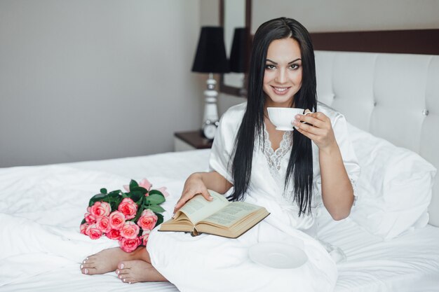 젊고 아름다운 브루네트 행복한 소녀는 장미 꽃다발을 들고 침대에 앉아 커피를 마시고 아침에 책을 읽습니다.
