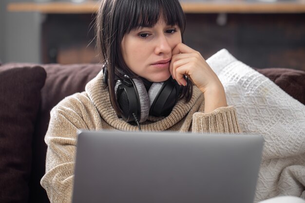 Young beautiful brunette in headphones in a cozy sweater and looks at the laptop screen.