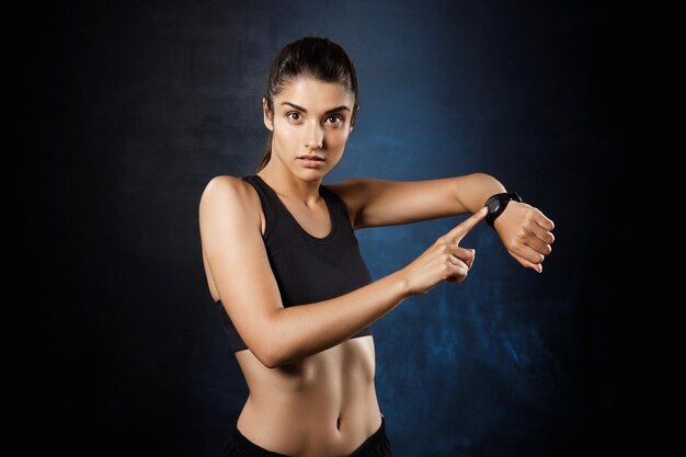 Young beautiful brunette sportive woman pointing at watch over dark wall