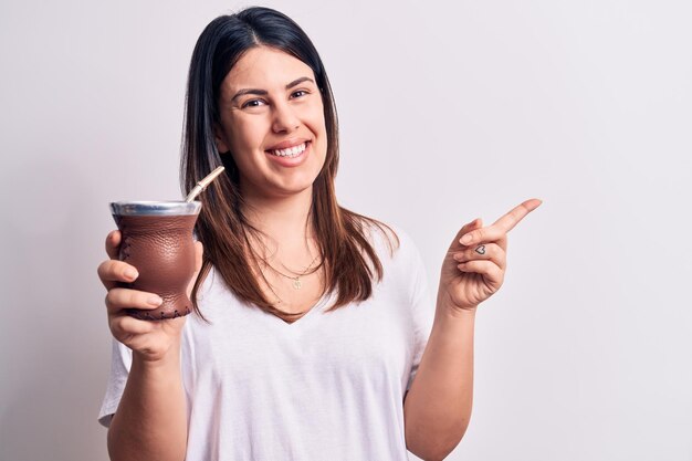 Free photo young beautiful brunette woman drinking mate infusion beverage over white background smiling happy pointing with hand and finger to the side