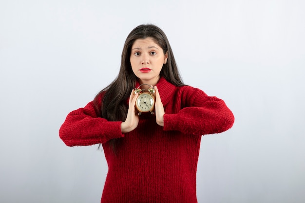 Young beautiful brunette woman holding an alarm clock standing over white background