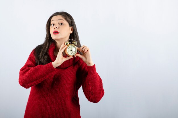 Young beautiful brunette woman holding an alarm clock standing over white background