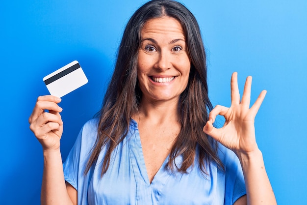 Young beautiful brunette woman holding credit card over isolated blue background doing ok sign with fingers smiling friendly gesturing excellent symbol