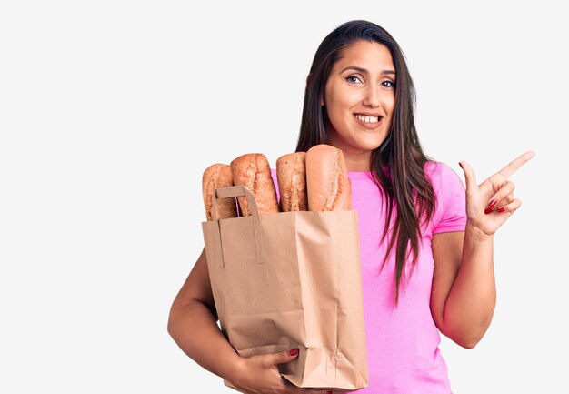 Молодая красивая брюнетка, держащая сумку для доставки с хлебом, счастливо улыбаясь, указывая рукой и пальцем в сторону