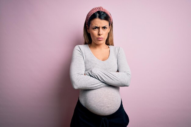 Молодая красивая брюнетка, беременная, ожидающая ребенка на изолированном розовом фоне, скептически и нервно неодобрительно смотрит на лицо со скрещенными руками.