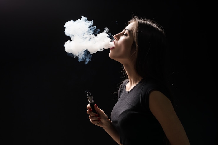 Free Photo young beautiful brunette woman smoking, vaping e-cigarette with smoke on the black wall