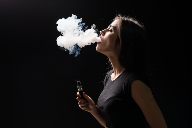 若い美しいブルネットの女性の喫煙、黒い壁に煙が電子タバコをvaping
